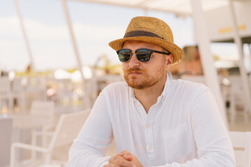 Smiling Man in White Shirt and Straw Hat Wearing Sunglasses at Outdoor Seaside Cafe