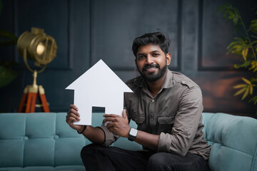 Smiling young Indian guy sitting on sofa and holding house, House loan and Reals state business...