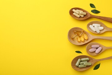 Different vitamin pills in spoons and green leaves on yellow background, flat lay. Space for text