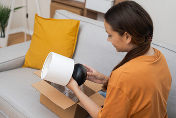 Woman takes out small table lamp from cardboard parcel with home delivery online purchase from...