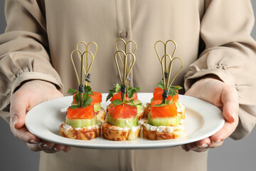 Woman holding tasty canapes with salmon, cucumber, bread and cream cheese on grey background,...