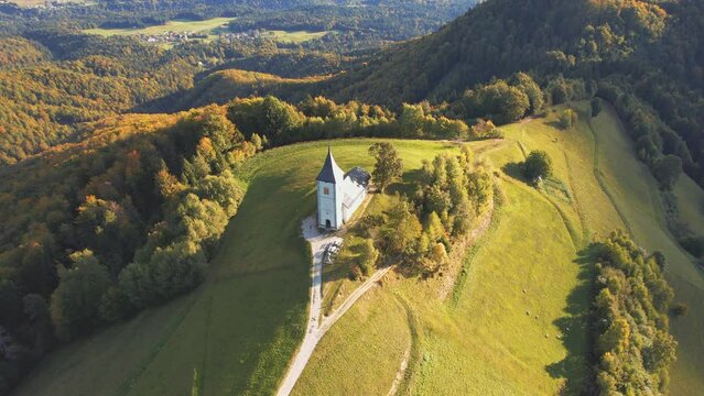Bird's eye (overhead) view with 360-degree video and tracking shot of a colourful autumn sunrise over the small catholic church of St. Primoz in Jamnik on a green hill in Slovenia
