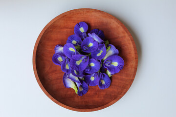 Obraz na płótnie Canvas Purple flower of Clitoria ternatea, or butterfly pea flower or bunga telang, on wooden plate, isolated on white background, flat lay or top view