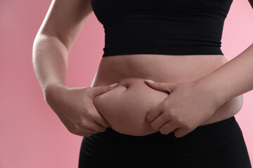 Woman touching belly fat on pink background, closeup. Overweight problem