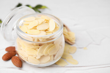 Fresh almond flakes in jar and nuts on white textured table, closeup. Space for text