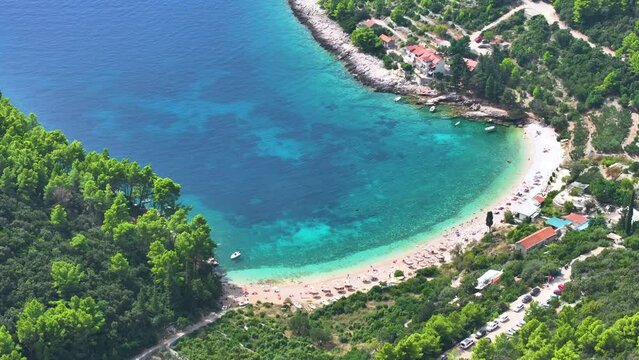 AERIAL: Flying above a scenic public beach with gorgeous turquoise water filled with tourists' umbrellas. Picturesque aerial view of a scenic public beach on Korcula island on a sunny summer day.