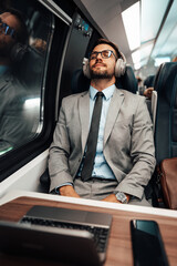 Tired handsome businessman relaxing while traveling with high-speed train or metro. He is using laptop computer and wireless headphones for entertainment and music listening.