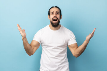 What do you want? Portrait of angry confused man standing with raised hands and surprised indignant expression, asking why how, what reason. Indoor studio shot isolated on blue background.