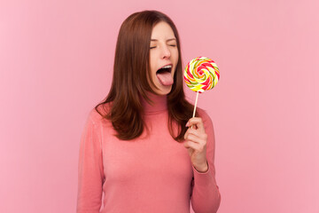 Hungry woman with brown hair sticking out tongue licking lollipop, tasting sweet round candy,...