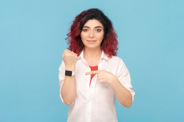Portrait of serious attractive woman with fancy red hair standing pointing at wrist watch,...