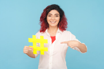 Portrait of attractive optimistic woman with fancy red hair holding pointing yellow hashtag,...