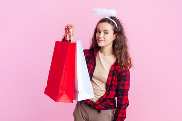 Portrait of angelic teenage girl with wavy hair in red checkered shirt and nimb over head standing showing paper shopping bags. Indoor studio shot isolated on pink background.