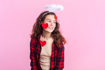Portrait of smiling cheerful teenage girl with wavy hair in red checkered shirt standing with nimb over head and coveredwith red paper hearts. Indoor studio shot isolated on pink background.