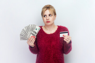Portrait of confused young blonde woman standing holding dollar banknotes and credit card, banking...