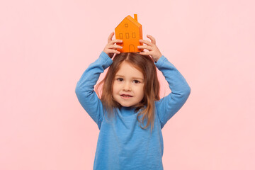 Portrait of adorable positive little girl raised her arms and holding paper house above her house,...