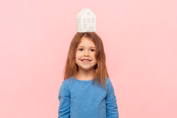 Portrait of delighted little girl with paper house on her head looking at camera, real estate...