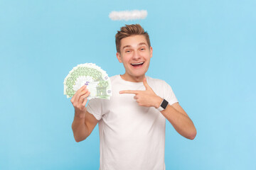 Portrait of rich angelic positive man wearing white t-shirt with nimb over head holding pointing at euro banknotes, getting salary. Indoor studio shot isolated on blue background