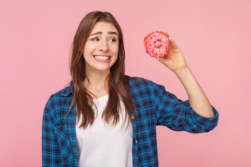 Portrait of funny brown haired woman holding donut looking away thinking eat or not keeps diet clenched teeth, wearing checkered shirt. Indoor studio shot isolated on pink background.