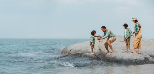 Asian family climbing rocks on the beach, father giving his hand to help son to climb down the...