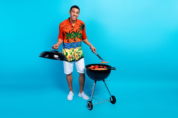 Full length photo of funky cheerful guy dressed print shirt grilling sausages isolated blue color...