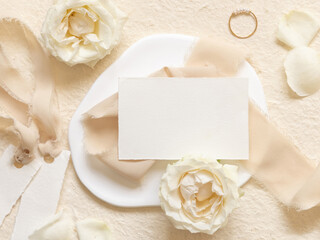 Card near cream roses and petals, ring and silk ribbons on beige top view, wedding mockup