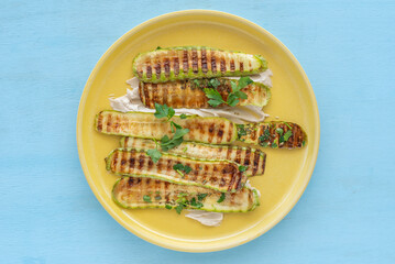 Grilled zucchini served with tahini pasta and chopped herbs on yellow platter over wooden...