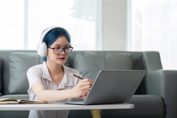 Smiling girl student wear headphone study online with teacher, happy young asian woman learn lecture watch webinar write notes look at laptop sit in home, distant education