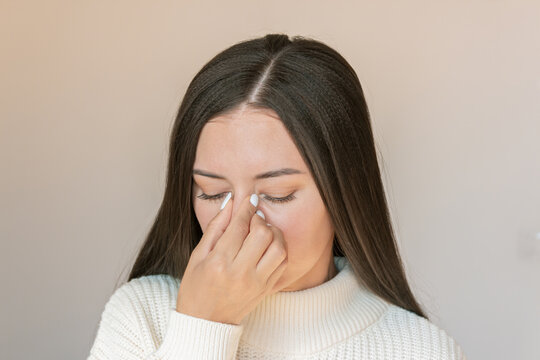 Young Caucasian woman suffers from nasal congestion and runny nose, experiences pain in the nose with sinusitis, holds her nose with her hand. Isolated on a beige background. Allergy