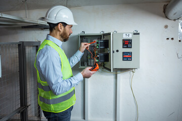 electrician in hardhat and safety vest holding tablet and fixing electric panel