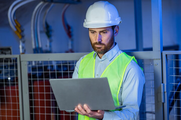 Caucasian factory worker in white hard hat and green safety vest typing on modern digital laptop at workplace.