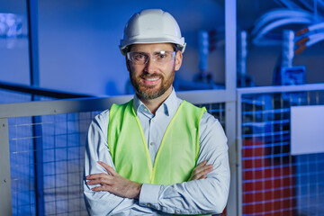 portrait of a handsome confident male caucasian engineer or worker wear safety hard hat, goggles, cross arms, smile among dark heavy industrial factory background