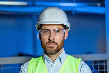 Handsome skilled bearded engineer in hardhat,goggles and vest looking at camera on blue background