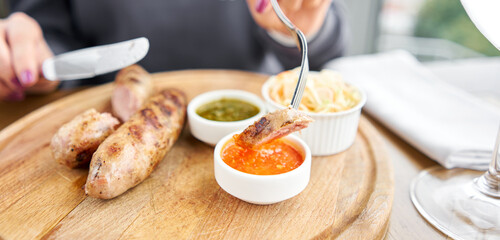 A piece of sausage is dipped in sauce. Lunch in a restaurant, a woman cuts Grilled sausages....