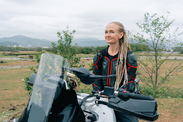 Female biker wearing Motorcycle Body Armor and dreadlocks standing near her motorcycle in moto trip in mountains