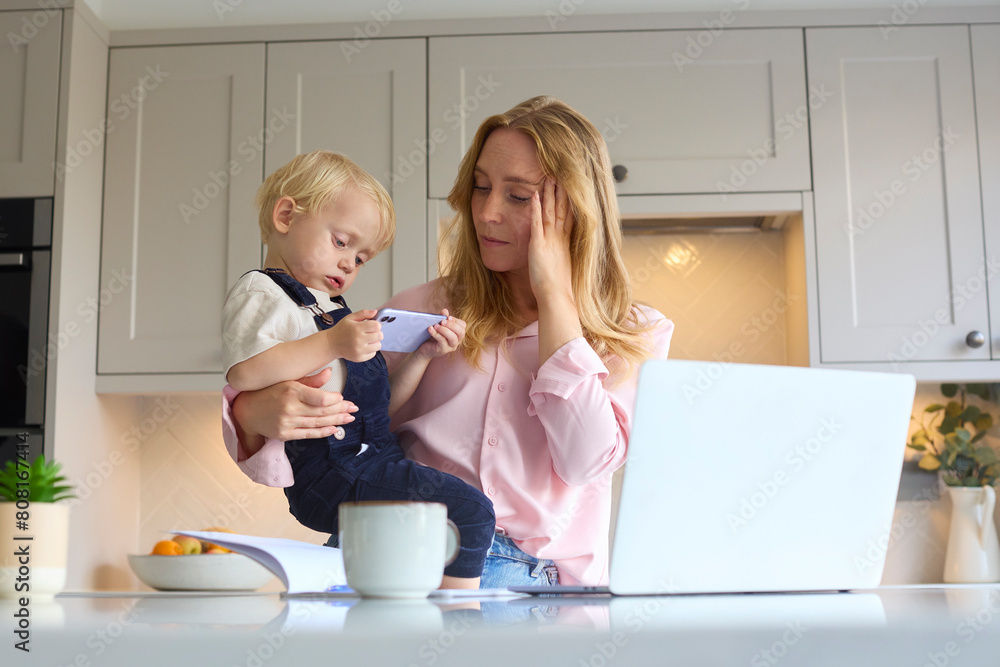 Sticker stressed working mother with young son at home in kitchen using laptop as boy plays with her phone