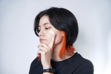 Mediterranean black and orange haired person applying a scoop of moisturizing cream on their cheek...