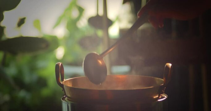 Slow motion scene of a boiling pot steams up as a restaurant's chef uses a ladle to stir soup in a cooking pot in the morning.