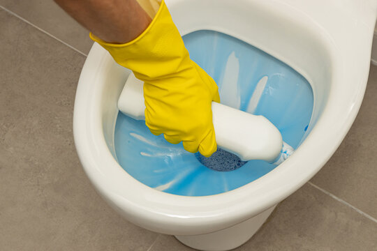 Close up of a hand wearing a protective yellow rubber glove applying blue liquid toilet cleaner under the rim of a white, modern, ceramic toilet bowl.