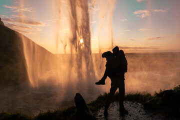 Couple kissing in silhouette under a huge waterfall. Hiker posing at sunset behind the...