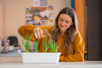 Woman gently waters her indoor garden of green onions, reflecting blend of home life and care for sustainable living, in her well-lit home office.