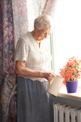 Sad old woman near the window. Happy senior woman sitting near window with flowers.