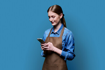 Young female worker in an apron using smartphone on blue studio background