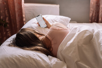 The girl feels bad and lies in bed holding a phone with a calendar in her hand.