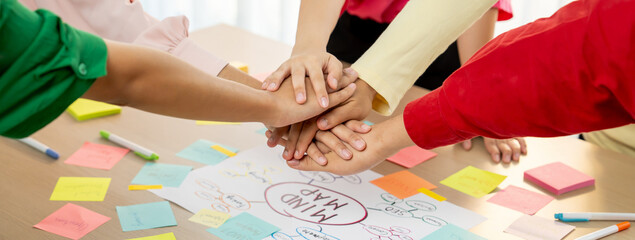 A group of business people putting their hands together at meeting room on table with mind map scatter around. Startup group showing unity teamwork and friendship. Close up. Focus on hand. Variegated.