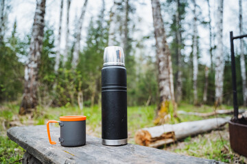 A thermos is on the table against the background of the forest, next to it is a tourist mug....