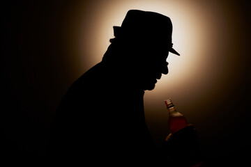 Silhouette of a man sat on his own drinking a bottle of whisky,with a glow in the background.