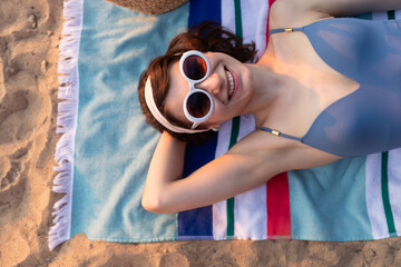 Young happy smiling woman in retro glasses and blue swimsuit lying on the beach striped towel. Summer vacation fashion and beauty lifestyle concept