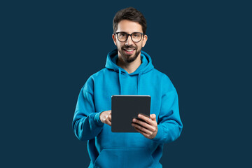A man wearing a blue hoodie is holding a tablet device in his hands. The man appears focused while interacting with the screen of the tablet. © Prostock-studio