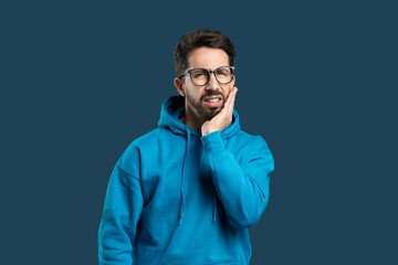 A man wearing glasses and a blue hoodie is standing against a neutral blue backdrop, holding his...