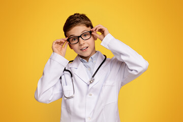 Pharmacy occupation. Friendly teen boy in doctor uniform posing, orange studio background